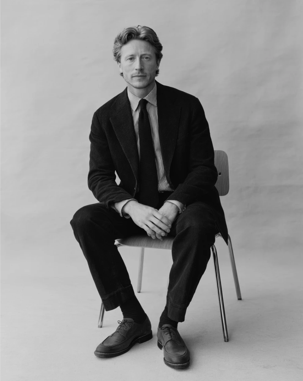 Black-and-white studio portrait of a man seated confidently on a chair in a formal suit, facing the camera against a plain backdrop, conveying professionalism and stability.